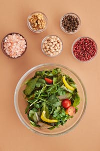 A vibrant salad with fresh greens, peppers, and tomatoes alongside bowls of spices and nuts, captured from above.