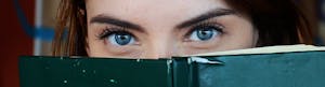 Close-up of a woman with blue eyes peeking over a book, portraying curiosity.