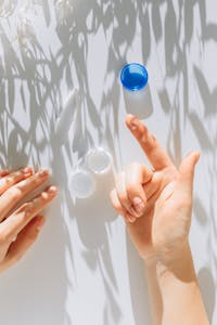 Close-up of hands holding a contact lens near a container, lit by soft sunlight on a white surface.