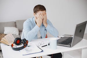 Stressed man at home office desk holding head in frustration over laptop work.