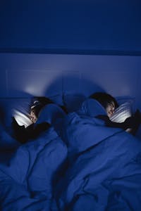 Two people lying in bed under blue light using smartphones, creating a cozy night mood.